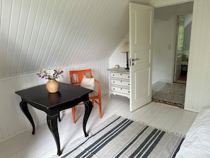 A room with a sloped ceiling, a wooden table with a vase, a chair, a dresser, and a bed.