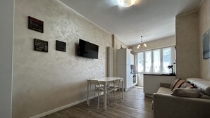 A modern living room with a sofa, a dining table with chairs, a flat-screen TV mounted on the wall, and a kitchen area visible through an open doorway. - [50m from the Beach] Modern Studio with WiFi & A/C (Lido Di fermo)