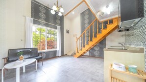 A spacious living area with a black leather sofa, a white coffee table, and a large window offering a view of trees. A wooden staircase with a patterned railing leads to a kitchen area with a sink and a tiled backsplash. - Yangpyeong Dodam Dodam Pension (Yangpyeong)