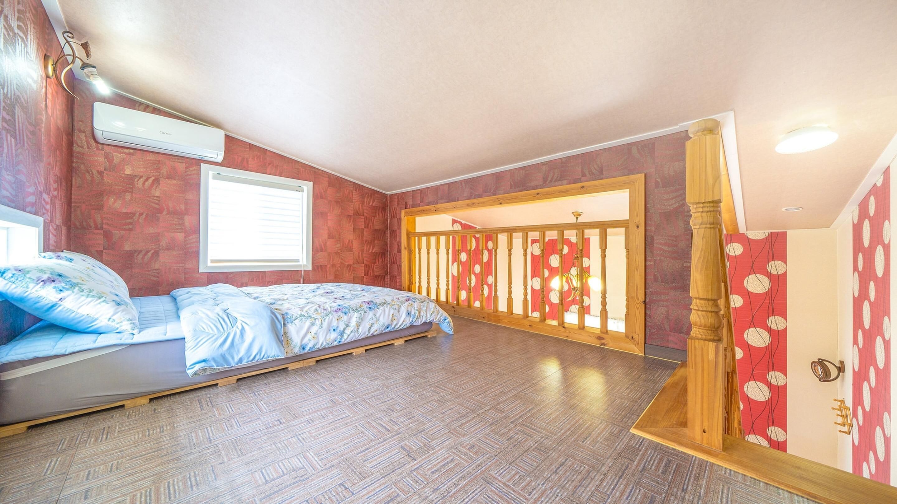 Un dormitorio con piso de madera, una cama con cubrecama floral, una ventana con persianas y una baranda de madera con diseño rojo y blanco.
