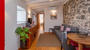A modern living room with a stone wall, a grey sofa, a wooden dining table, and a kitchen area with wooden cabinets and a potted plant. - Palace Guest Inn (Queluz)
