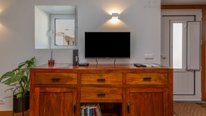 A living room with a flat-screen TV mounted on a wooden cabinet, a wall-mounted light, and a potted plant. - Palace Guest Inn (Queluz)
