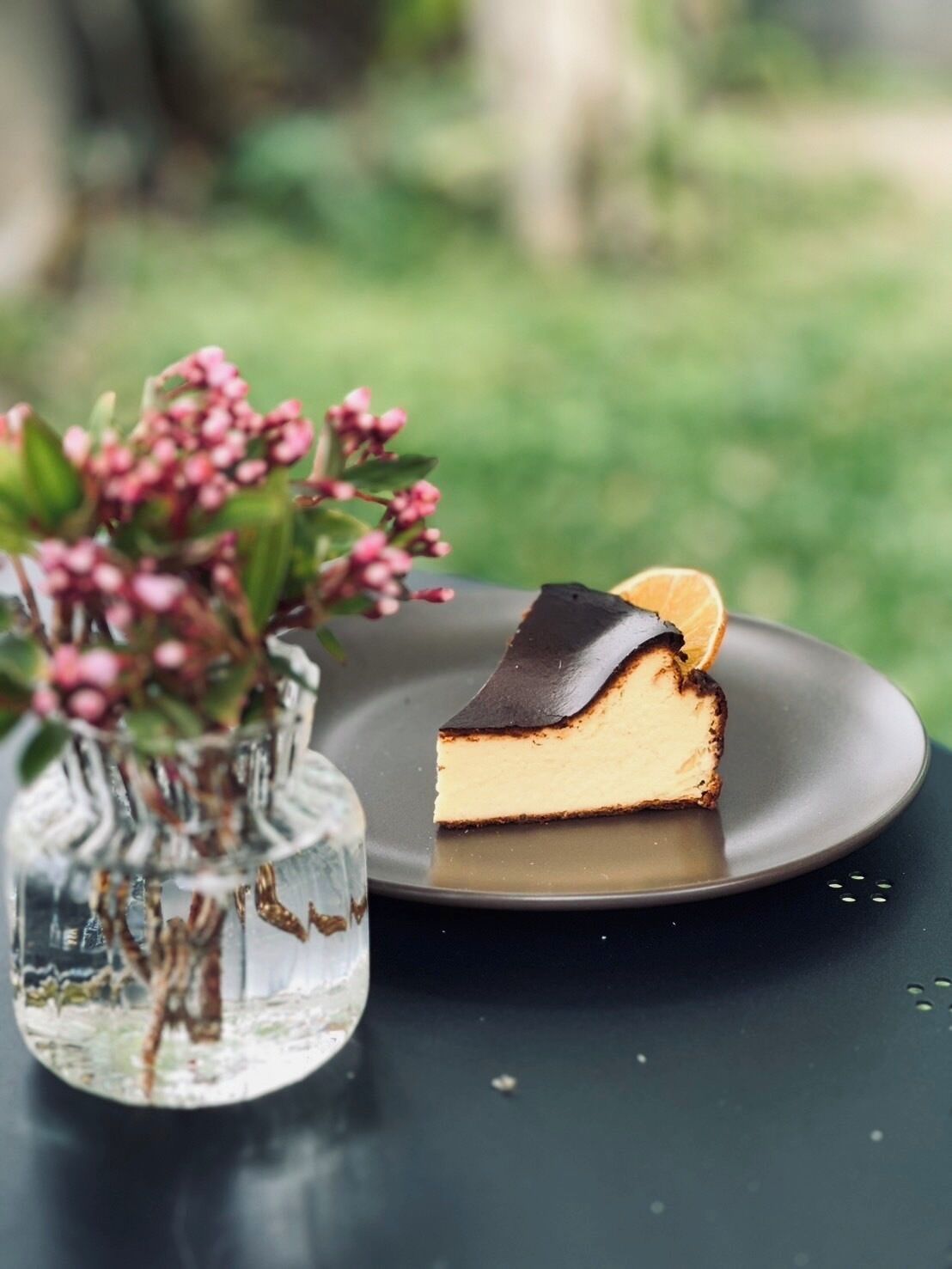 A slice of cheesecake on a dark plate with a dark chocolate topping and an orange slice on top, placed on a reflective surface.