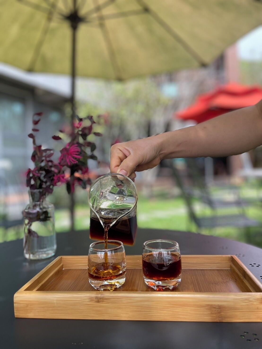 A hand pouring a dark liquid into two glasses on a wooden tray.