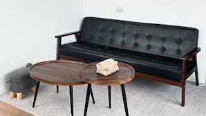 Un canapé en cuir noir avec une structure en bois, deux petites tables rondes en bois et un petit pouf gris.