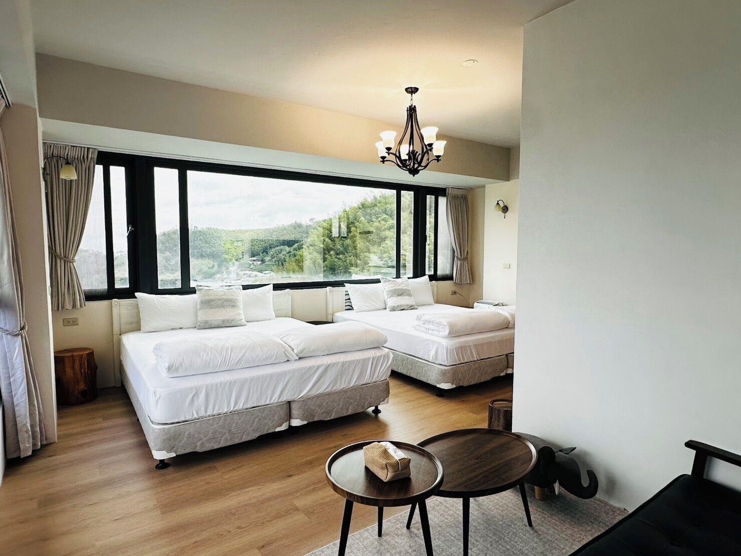 A bedroom with two beds, a chandelier, a view of greenery, and a small round table.