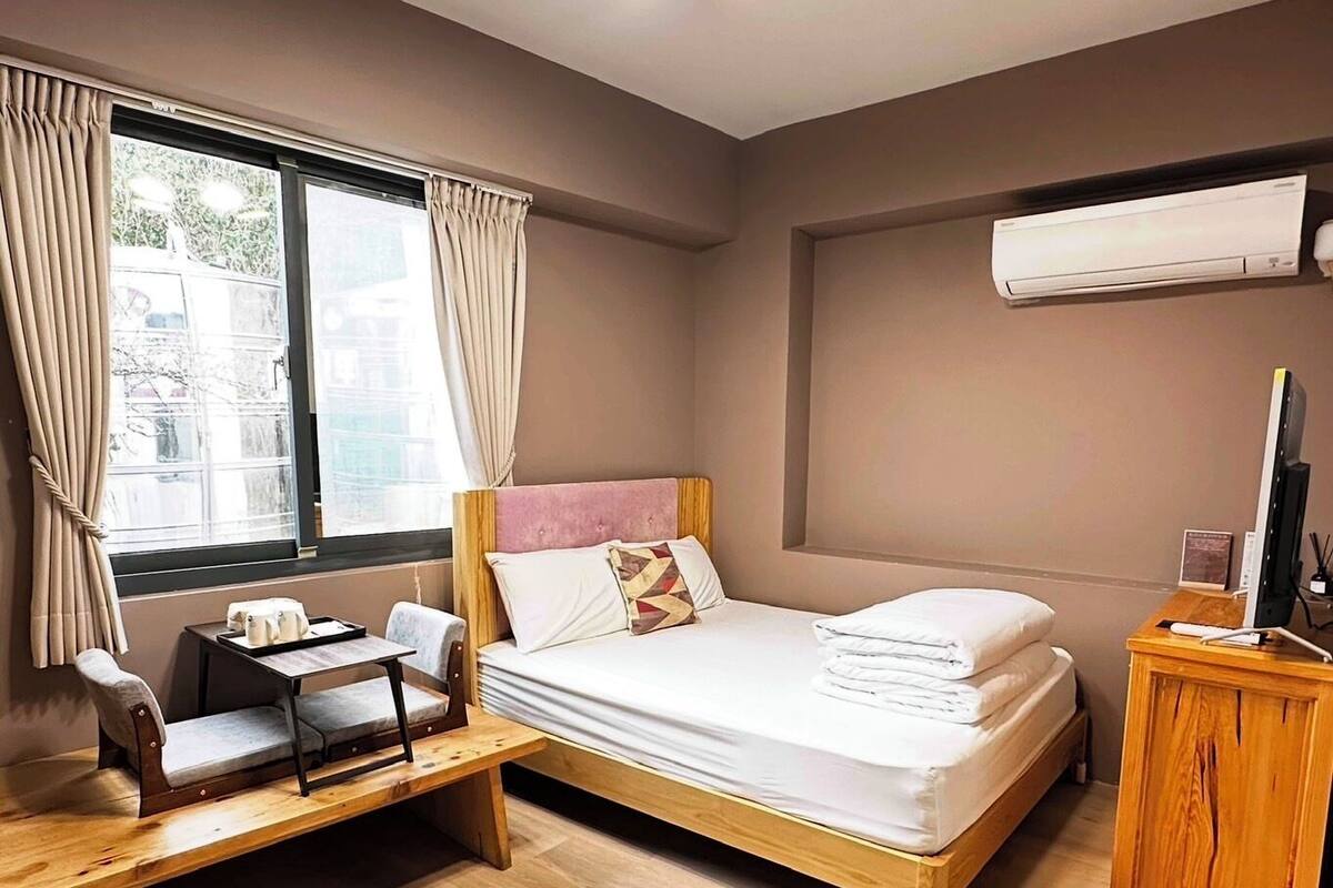 A hotel room with a bed, a desk, a chair, an air conditioning unit, and a window with curtains.