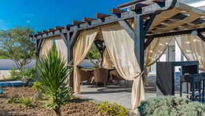 Un espace extérieur aménagé en terrasse, doté d’une pergola recouverte de rideaux beiges et agrémenté de tables et de chaises en osier.