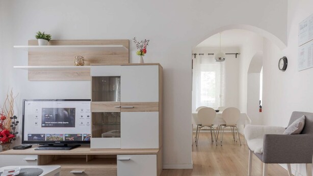 Ein modernes Wohnzimmer mit integriertem Fernseher, einem Essbereich mit weißen Stühlen und einem grauen Sofa.