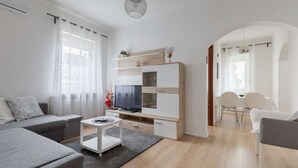 Ein modernes Wohnzimmer mit grauem Sofa, einem weißen Couchtisch, einem Fernseher auf einer Holzkommode und einem Essbereich mit weißen Stühlen.