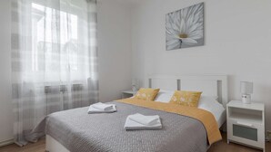 Ein ordentlich gemachtes Bett mit einer grauen und gelben Tagesdecke, weißen Kissen und gefalteten Handtüchern. Ein Nachttisch mit Lampe und ein kleiner, weißer Beistelltisch ebenfalls mit Lampe. Ein großes Fenster mit transparenten Vorhängen lässt natürliches Licht in den Raum.