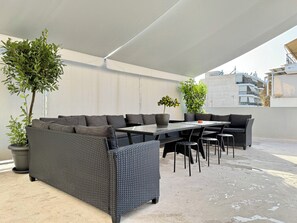 A covered outdoor seating area with a large woven sofa, a dining table, and several chairs. There are potted plants and a view of buildings in the background. - P7 - 2 Bedrooms Jacuzzi Rooftop (Athens)