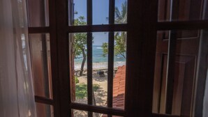 A view through a window with metal bars, revealing a beach with palm trees, a sandy area, and a red-roofed structure. - South Point Abbey Whole Villa 4 Bedroom (Sleeps 9) (Koggala)