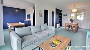 Ein modernes Wohnzimmer mit grauem Sofa, Couchtisch aus Holz und einem Essbereich mit Holztisch und Stühlen. Ein Fenster bietet Blick auf Bäume, und eine Pendelleuchte ist angebracht.