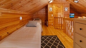 Ein gemütliches Schlafzimmer im Dachgeschoss mit Holzwänden und schrägem Dach. Es beinhaltet ein Bett, eine Kommode und einen Fernseher auf einem Holzboden.