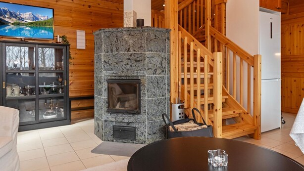 Ein gemütliches Blockhaus-Interieur mit Kamin aus Stein, einer Holztreppe und einem Fernseher, der eine Berglandschaft zeigt.
