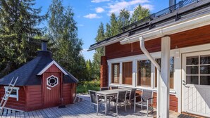 Eine Holzterrasse mit Esstisch und Stühlen, ein kleines rotes Gebäude mit Schornstein sowie eine weiße Tür mit Fenstern.