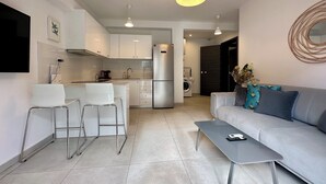 A modern kitchen with white cabinets, a built-in oven, and a stainless steel refrigerator. There is a dining area with a white table and chairs, and a gray sofa with decorative pillows. - P9 - 4 Bedroom apartments - Jacuzzi rooftop (Athens)
