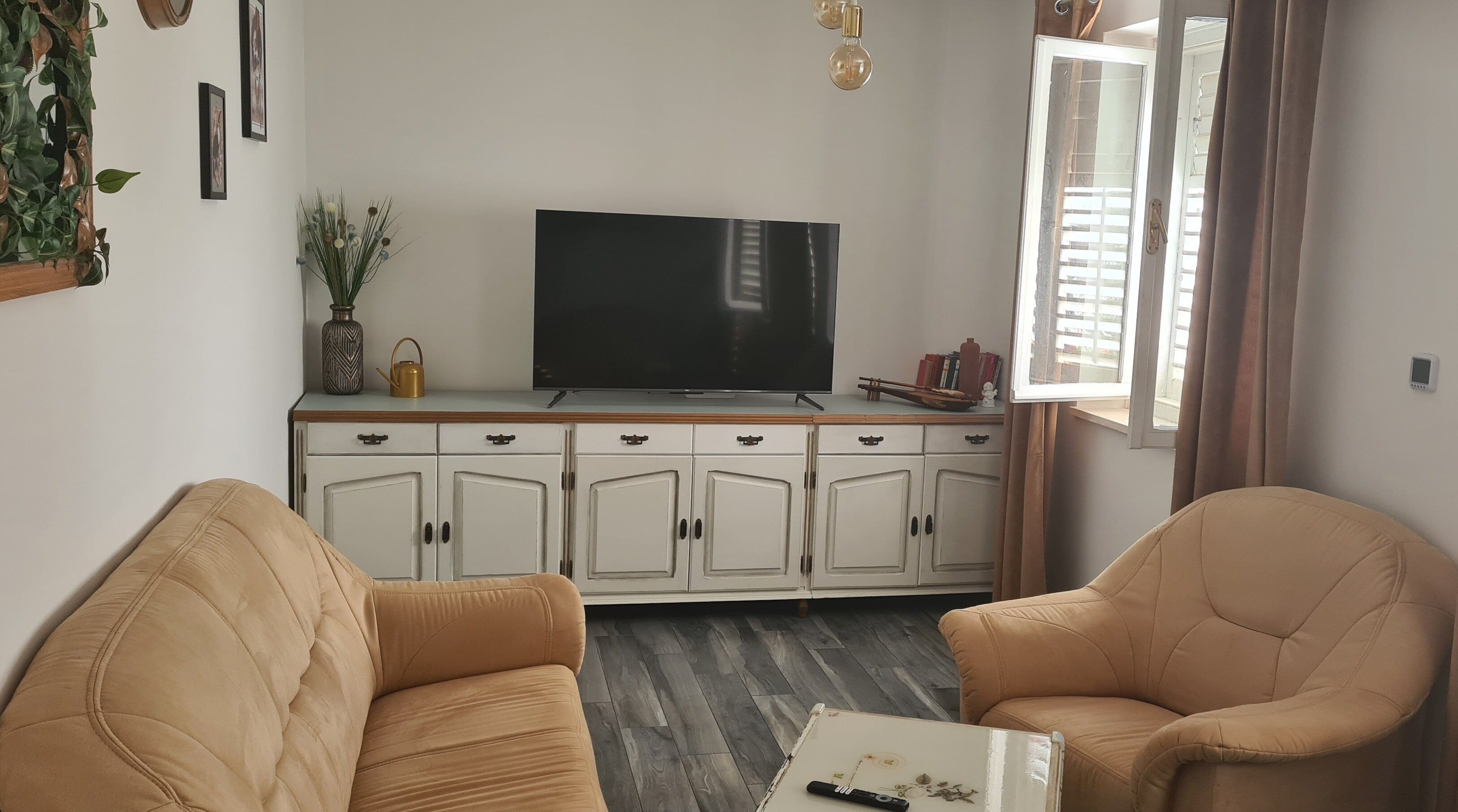 A living room with a flat-screen TV on a wooden cabinet, two beige sofas, a small table with a remote, and a window with curtains.