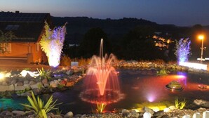 Área externa noturna com uma fonte de água, plantas iluminadas e um painel solar em um edifício.