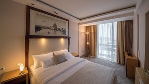 A hotel room with a large bed, a desk, a lamp, a picture on the wall, and a window with curtains. - Putnik Inn Belgrade (Belgrade)