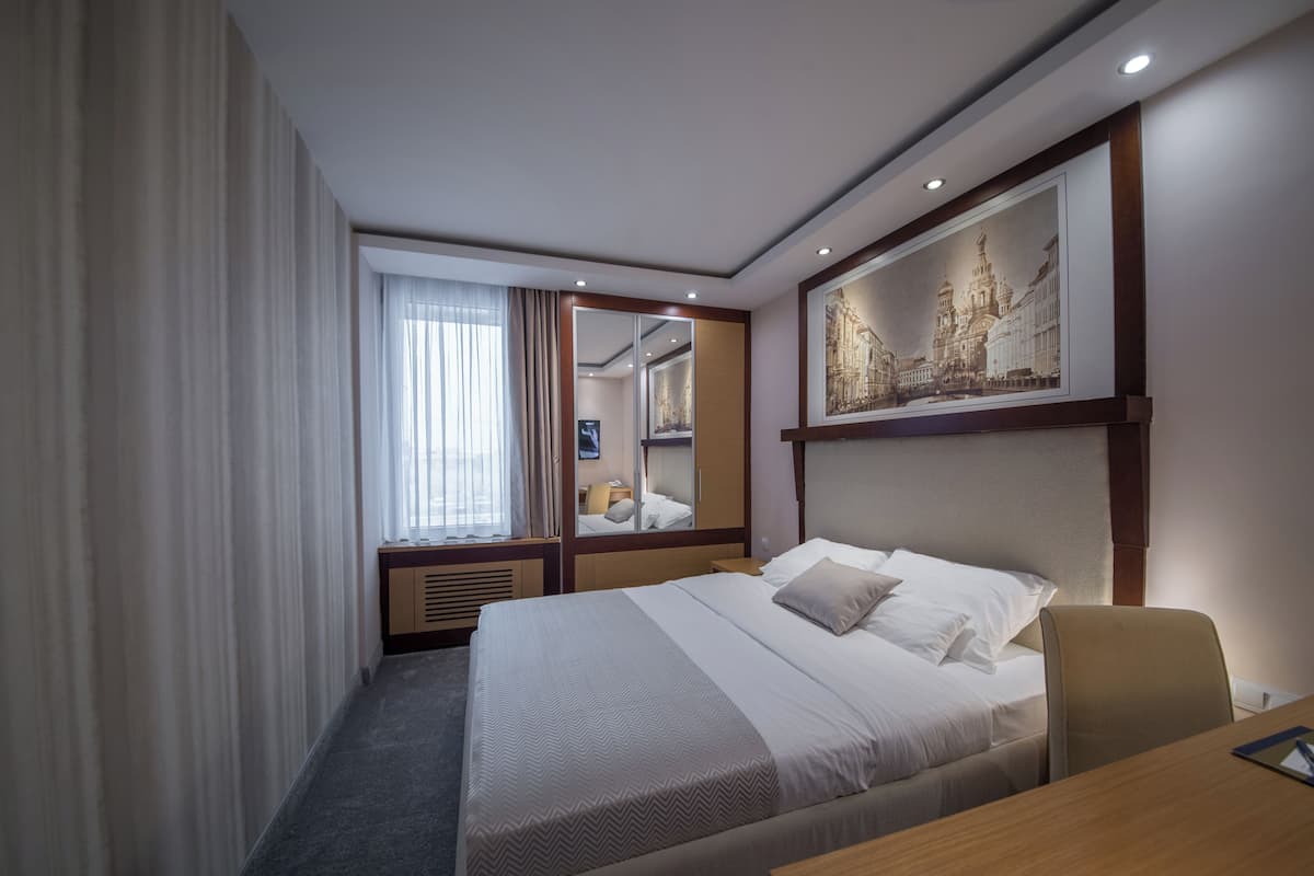 Ein Hotelzimmer mit einem großen Bett, einem Schreibtisch, einem Stuhl, einem Fenster mit Vorhängen, einem Spiegel und einem gerahmten Bild an der Wand.