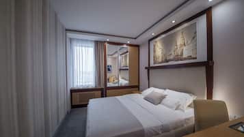 Ein Hotelzimmer mit einem großen Bett, einem Schreibtisch, einem Stuhl, einem Fenster mit Vorhängen, einem Spiegel und einem gerahmten Bild an der Wand.