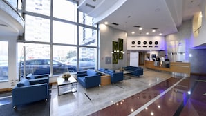 A modern hotel lobby with large windows, blue seating, a reception desk, and a visible parking area outside. - Putnik Inn Belgrade (Belgrade)