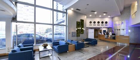 Un moderno vestíbulo de hotel con ventanas amplias, asientos azules, un mostrador de recepción y un estacionamiento visible en el exterior.