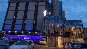 A hotel with a modern glass facade, a prominent sign, and a parking lot with several cars. - Putnik Inn Belgrade (Belgrade)