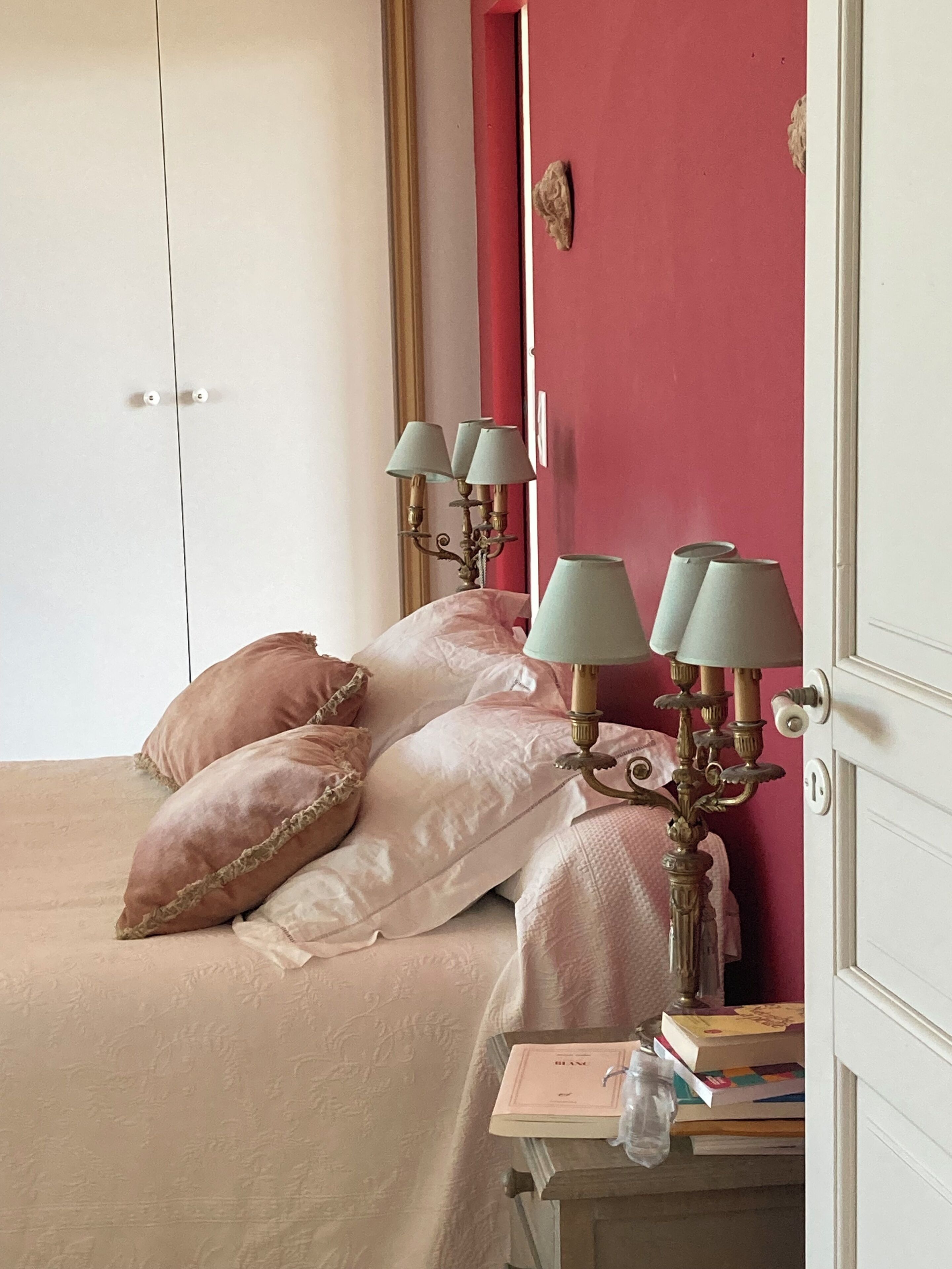 Un dormitorio con cama, almohadas, una mesita de noche con libros y una lámpara.