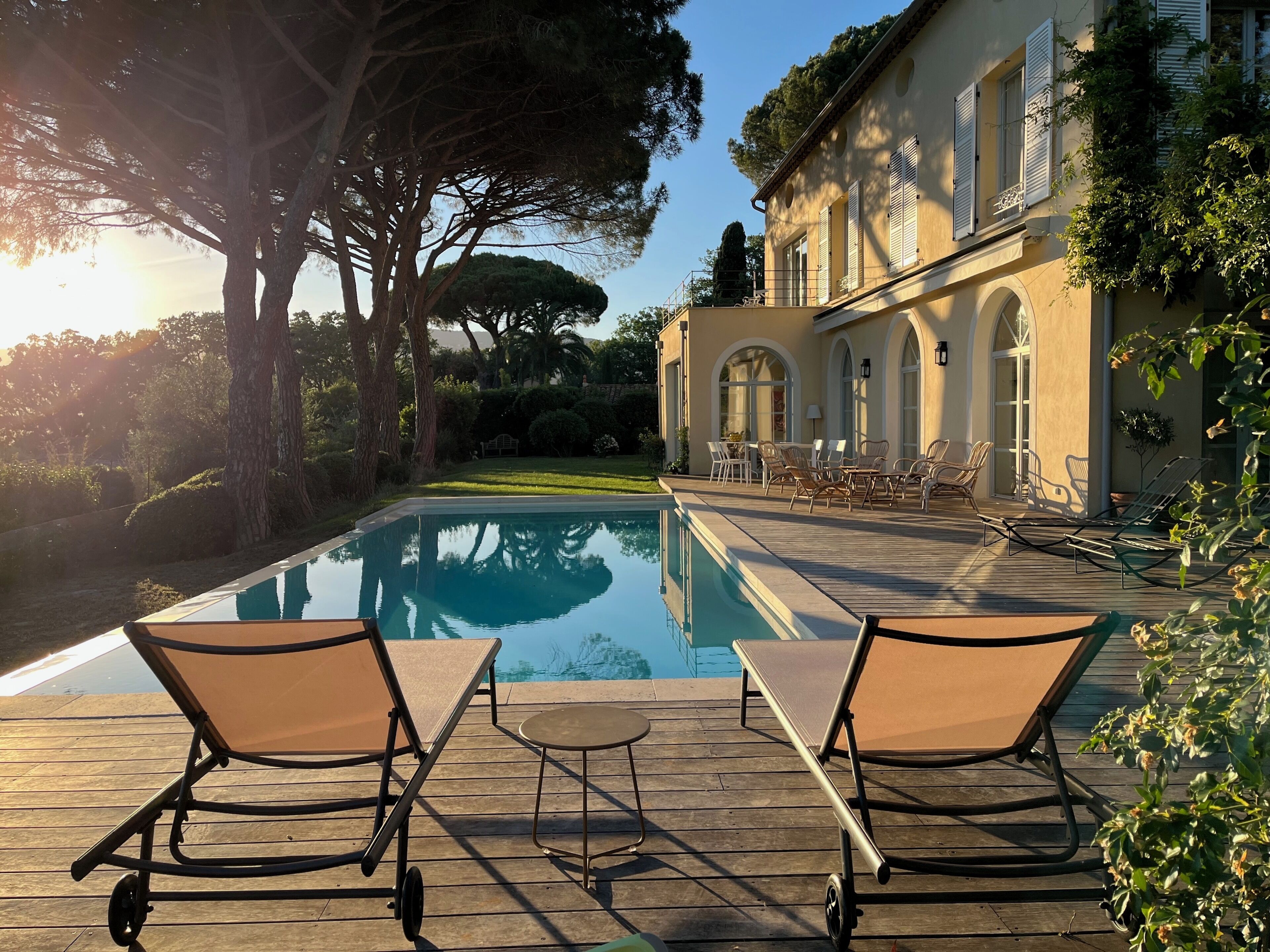 A pool with lounge chairs, a wooden deck, and a building with arched windows and shutters.