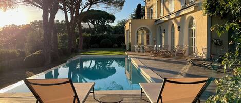 A pool with lounge chairs, a wooden deck, and a building with arched windows and shutters.