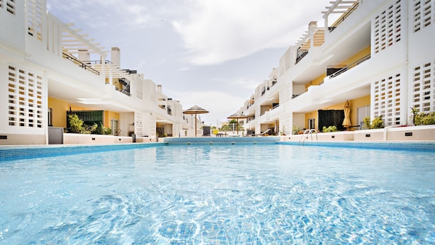 A swimming pool with clear blue water, surrounded by white buildings with balconies and outdoor seating areas.