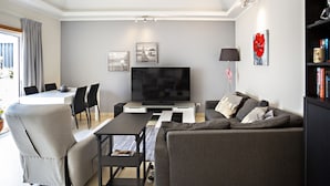 A modern living room with a flat-screen TV, grey sofa, dining table, chairs, and a bookshelf.