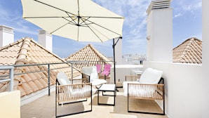 A rooftop terrace with white wicker furniture, a large umbrella, and a tiled roof.
