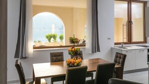 A dining area with a wooden table and chairs, a fruit basket, and a view of a body of water and a tower through a large window. - Apartment Frankie's Place (Dubrovnik)