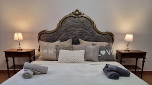 Cama bem arrumada com cabeceira, duas mesas de cabeceira com abajures e várias almofadas com texto.