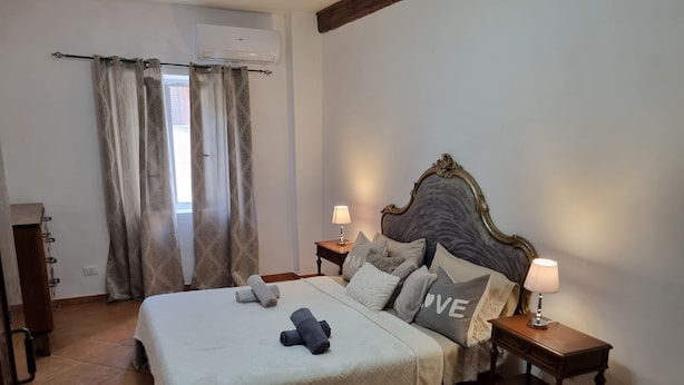 Um quarto com cama, almofadas, mesa de cabeceira, abajur, janela com cortinas e um ar condicionado.