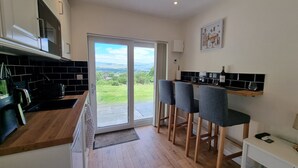 A modern kitchen with a sliding glass door leading to a balcony, wooden flooring, and bar stools. - Studio | Sleeps 3 | Mountain Views - 5 Mins from Kenmare town. (Kenmare)