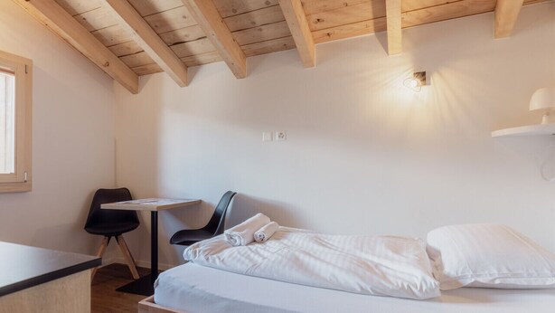 A room with a wooden ceiling, a bed with white bedding, a small table, a chair, and a window. - Studio Rénové à Grimentz (Grimentz)