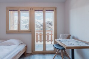 Une chambre comprenant un lit, une table avec une chaise et une porte-fenêtre coulissante donnant sur un balcon avec vue sur des montagnes enneigées.