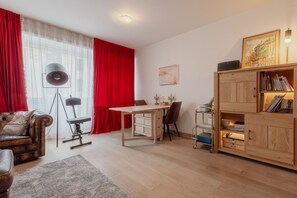 A living room with a wooden desk, a leather sofa, a bookshelf, and a large window with red curtains.