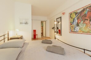 A hotel room with a bed, bedside tables, a mirror, a painting, and a hallway leading to a bathroom.