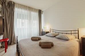 Un dormitorio con cama, almohadas, una mesita de noche, una lámpara y un banco.
