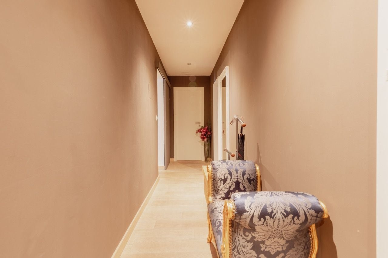 A hallway with a door at the end, a chair with a patterned upholstery, and a vase with flowers.