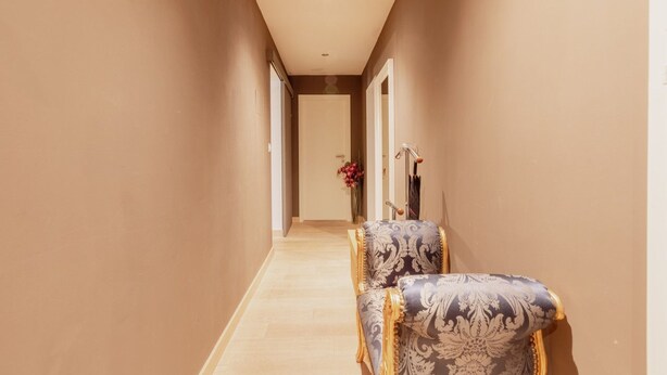 A hallway with a door at the end, a chair with a patterned upholstery, and a vase with flowers.