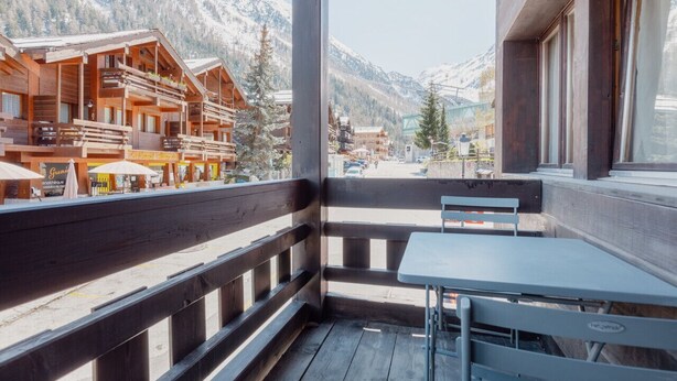 Un balcón de madera con una mesa y sillas, con vistas a un pueblo de montaña con edificios y picos nevados.