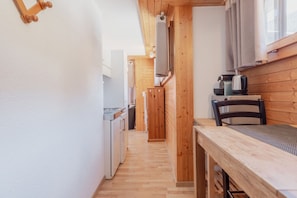 A narrow hallway with wooden walls and a wooden table with chairs. A kitchen area is visible in the background with appliances and a window.
