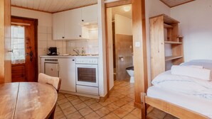 Une pièce compacte comprenant une table à manger en bois, un coin cuisine avec une cuisinière et un évier, un lit avec du linge de lit blanc et une étagère en bois.